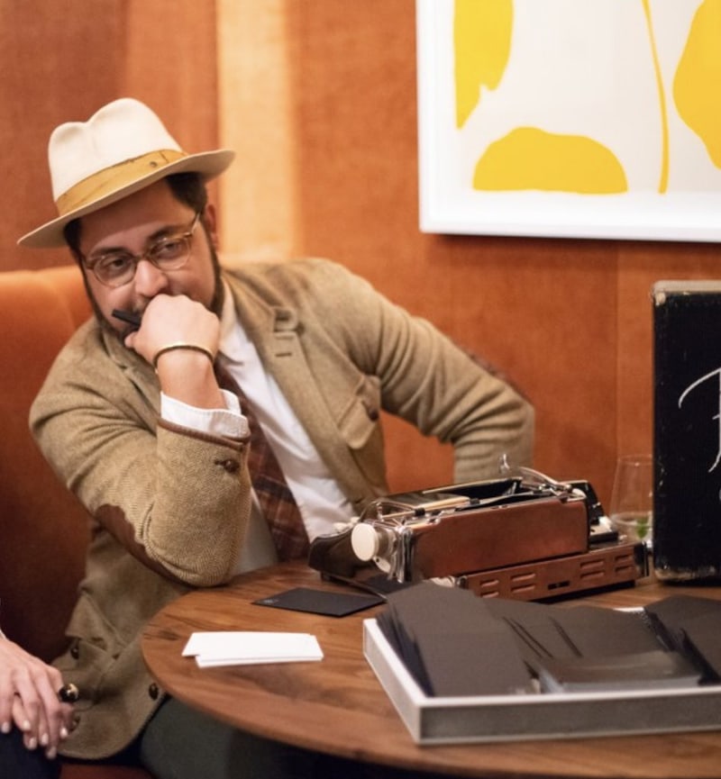 Paasha Motamedi in a cream fedora in an orange velvet banquette, chin on his hand, a chrome typewriter on the table