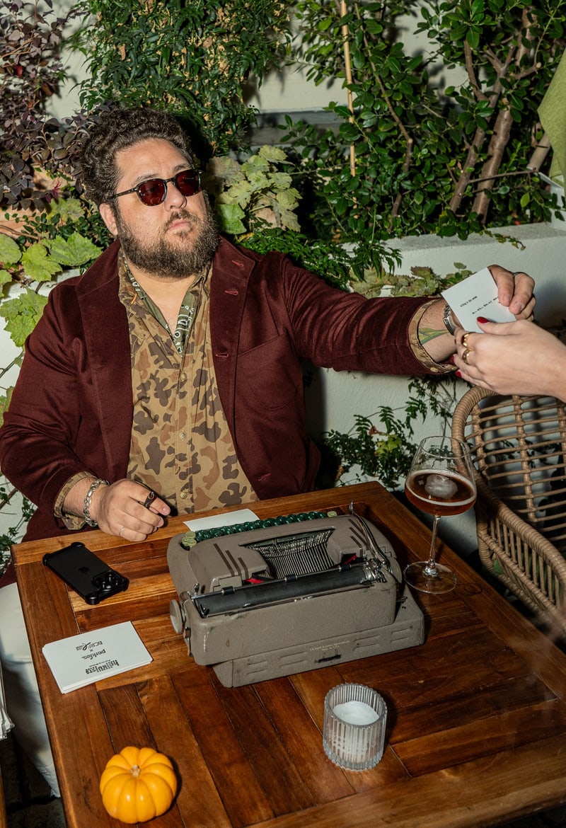 Paasha Motamedi in a burgundy velvet jacket handing a freshly typed poem across a garden table, a Smith-Corona typewriter in front of him
